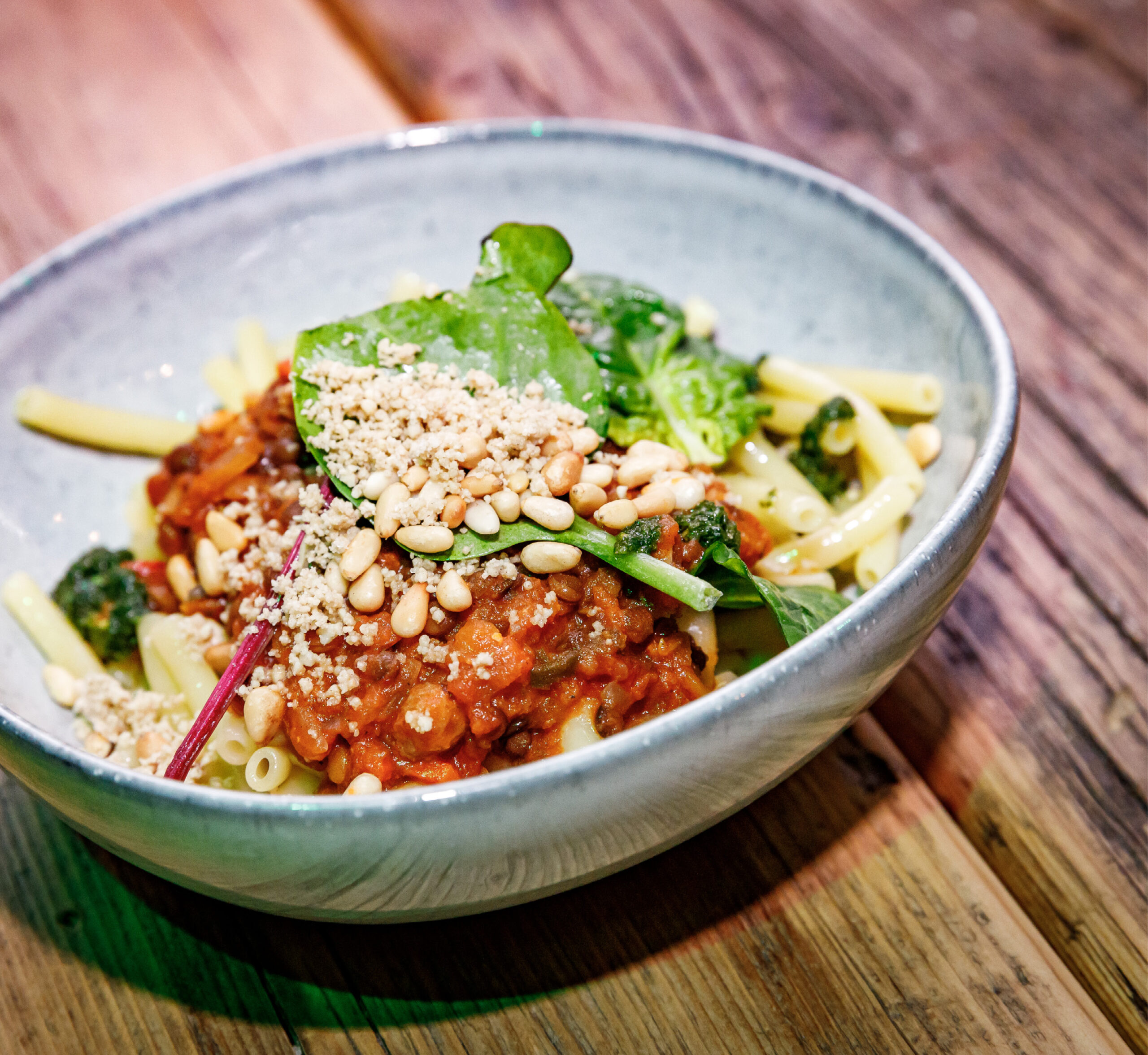 Eine Schüssel Nudeln mit Tomatensauce, Blattgemüse, Pinienkernen und geriebenem Käse auf einem Holztisch.