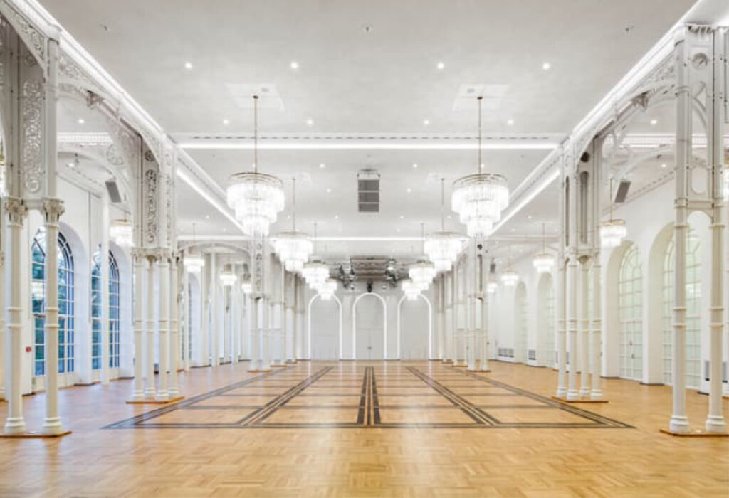Großzügiger, eleganter Ballsaal mit Kronleuchtern, Bogenfenstern, weißen Säulen und Holzparkettboden – perfekt für Veranstaltungen mit exquisitem Catering in der Flora Köln.