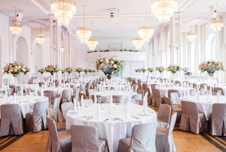 Eleganter Ballsaal mit runden Tischen, grauen Stühlen, floralen Tischdekorationen und Kronleuchtern für eine Veranstaltung – perfekt für Catering in der Flora Köln.