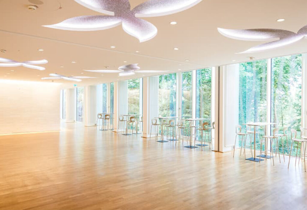 Großzügiger, moderner Raum mit großen Fenstern, Holzboden, Stehtischen, einzigartiger Deckenbeleuchtung – ideal für Veranstaltungen mit Catering in der Flora Köln.