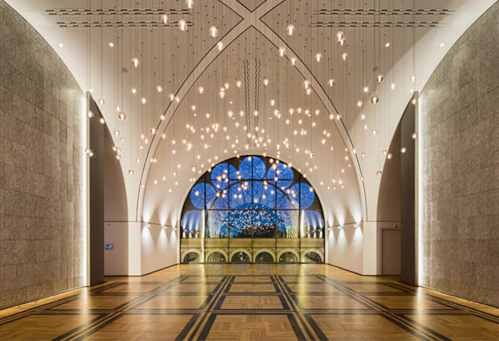 Großzügiger Saal mit gewölbter Decke, Hängelampen und einem großen Fenster mit blauem Ornamentglas – perfekt für Veranstaltungen mit Catering in der Flora Köln.