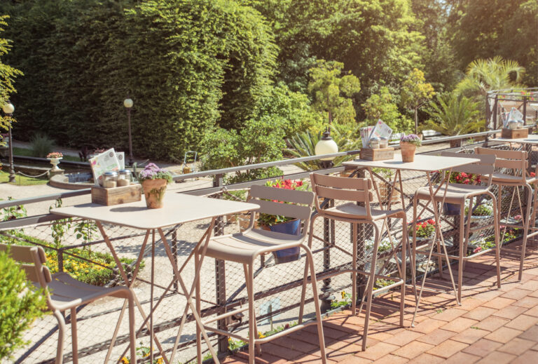 Café-Terrasse im Freien mit leeren Tischen und Stühlen und Blick auf einen üppigen grünen Garten an einem sonnigen Tag – perfekt zum Entspannen oder für ein Catering in der Flora Köln.