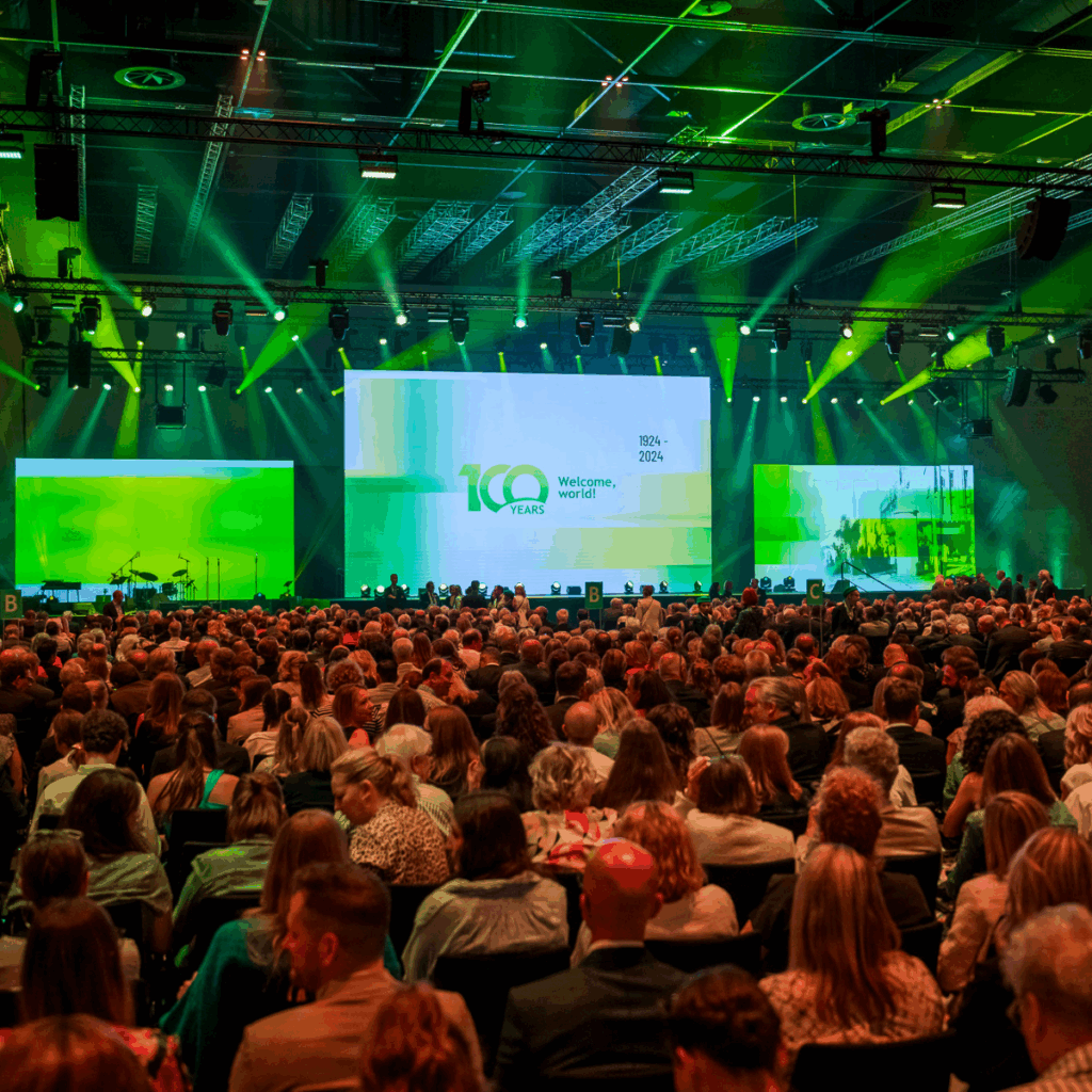 Großes Publikum sitzt in einem Saal mit Blick auf eine Bühne mit grüner Beleuchtung und einer „100 Jahre“-Ausstellung.