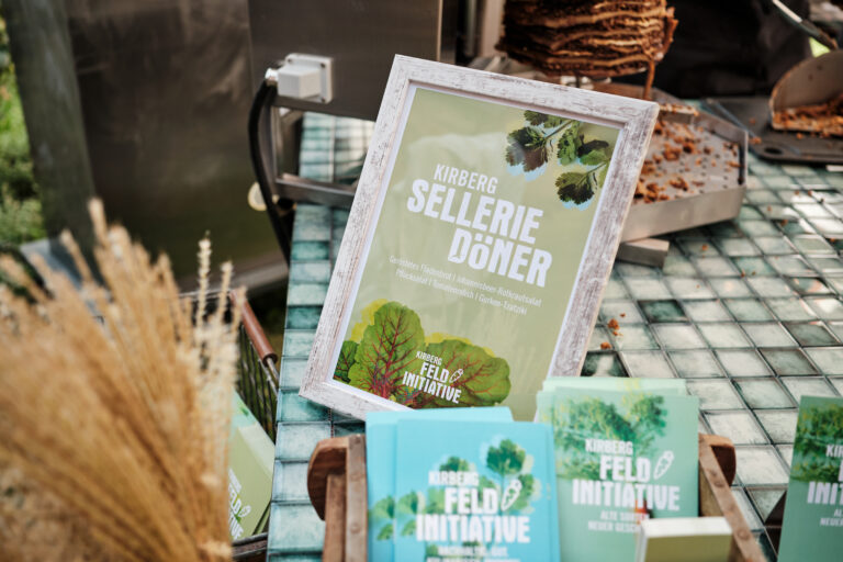 Gerahmtes Sellerie-Döner-Schild mit Blattgrafiken auf einem Tisch neben Büchern und einem vertikalen Drehspieß.