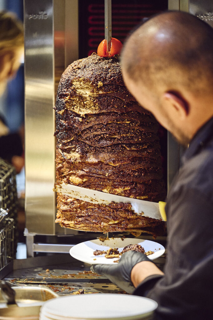 Ein Mann schneidet in einem Restaurant Fleisch von einem vertikalen Drehspieß auf einen Teller.