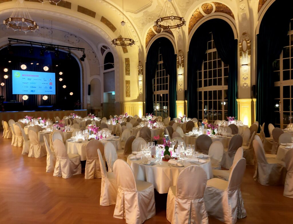 Eleganter Bankettsaal mit runden Tischen, weißen Stühlen, Blumengestecken und einer Bühne mit großer Leinwand.
