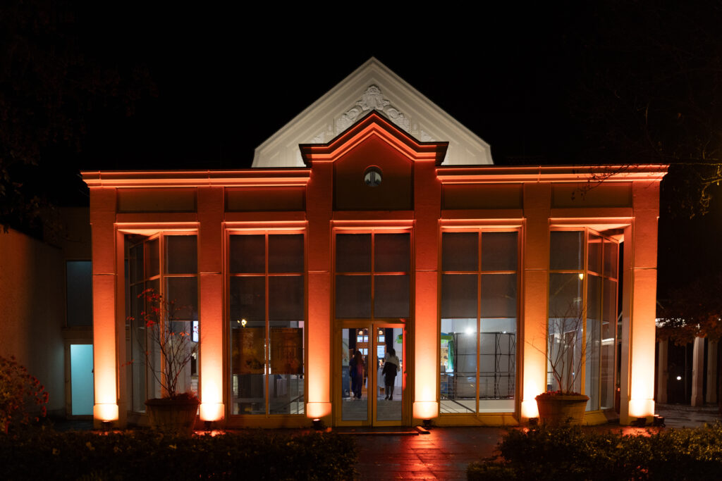 Kurhaus Bad Honnef - Ein Gebäude mit Glasfront, das nachts orangefarben beleuchtet wird. Durch den Eingang sind die Menschen im Gebäude sichtbar.