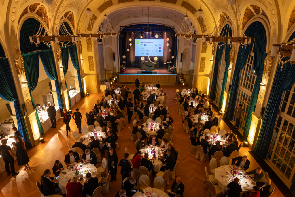 Ein formelles Bankett mit runden Tischen und Gästen in einem reich verzierten Saal vor einer Bühne mit einer großen Leinwand.