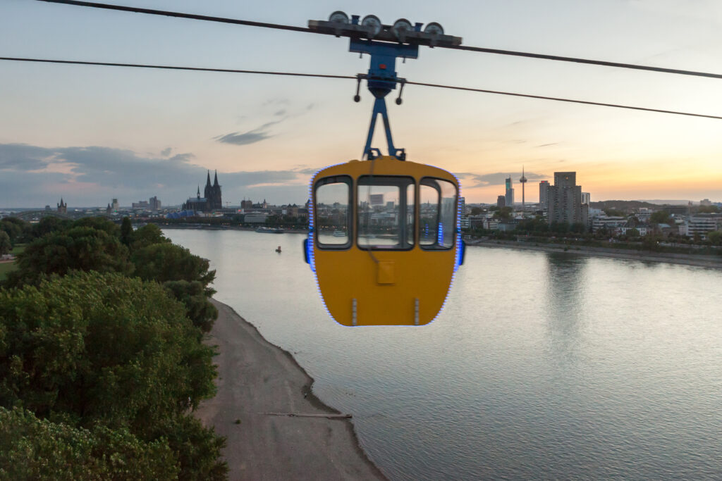 Eine gelbe Seilbahn überquert bei Sonnenuntergang einen Fluss, im Hintergrund die Skyline einer Stadt und eine Kathedrale.