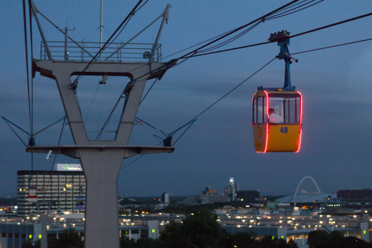 Eine gelbe Seilbahn mit roten Lichtern fährt in der Abenddämmerung über eine Stadtlandschaft mit Gebäuden und einem Stadion im Blick.