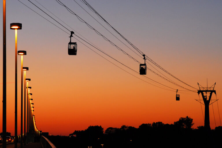 Seilbahnen fahren bei Sonnenuntergang auf Drähten und zeichnen sich als Silhouette vor einem orangefarbenen Himmel mit Bäumen darunter ab.