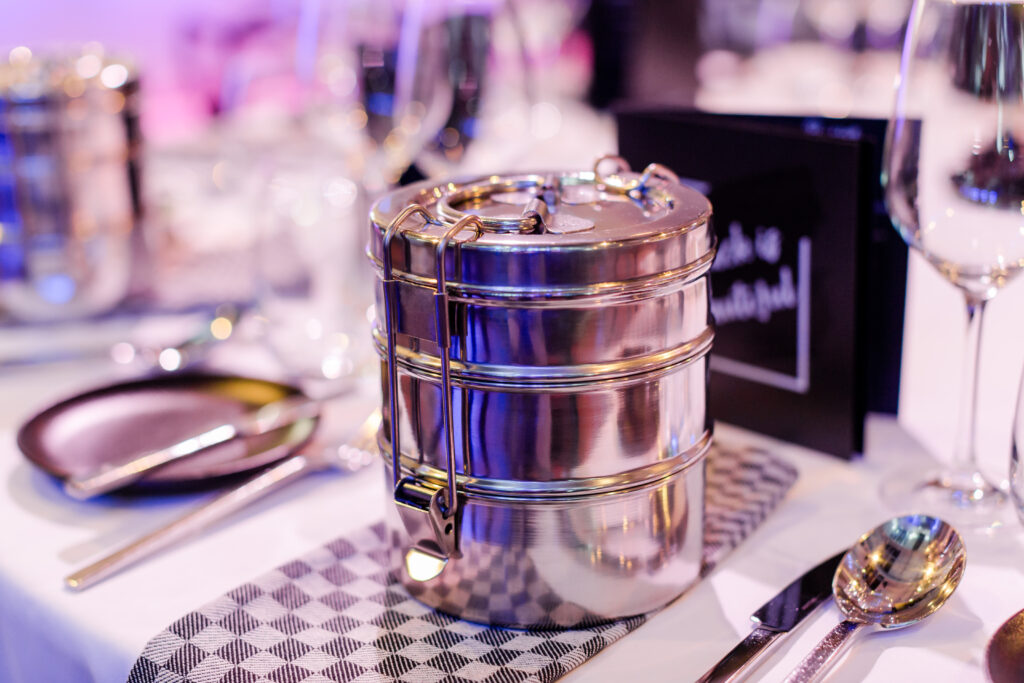 Eine Tiffin-Box aus Edelstahl steht auf einer Stoffserviette an einem gedeckten Esstisch mit Glaswaren.