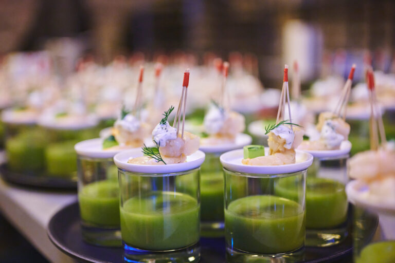 Kleine Gläser mit grüner Suppe, garniert mit aufgespießten Garnelen und Kräutern, ordentlich auf einem Serviertablett angerichtet.
