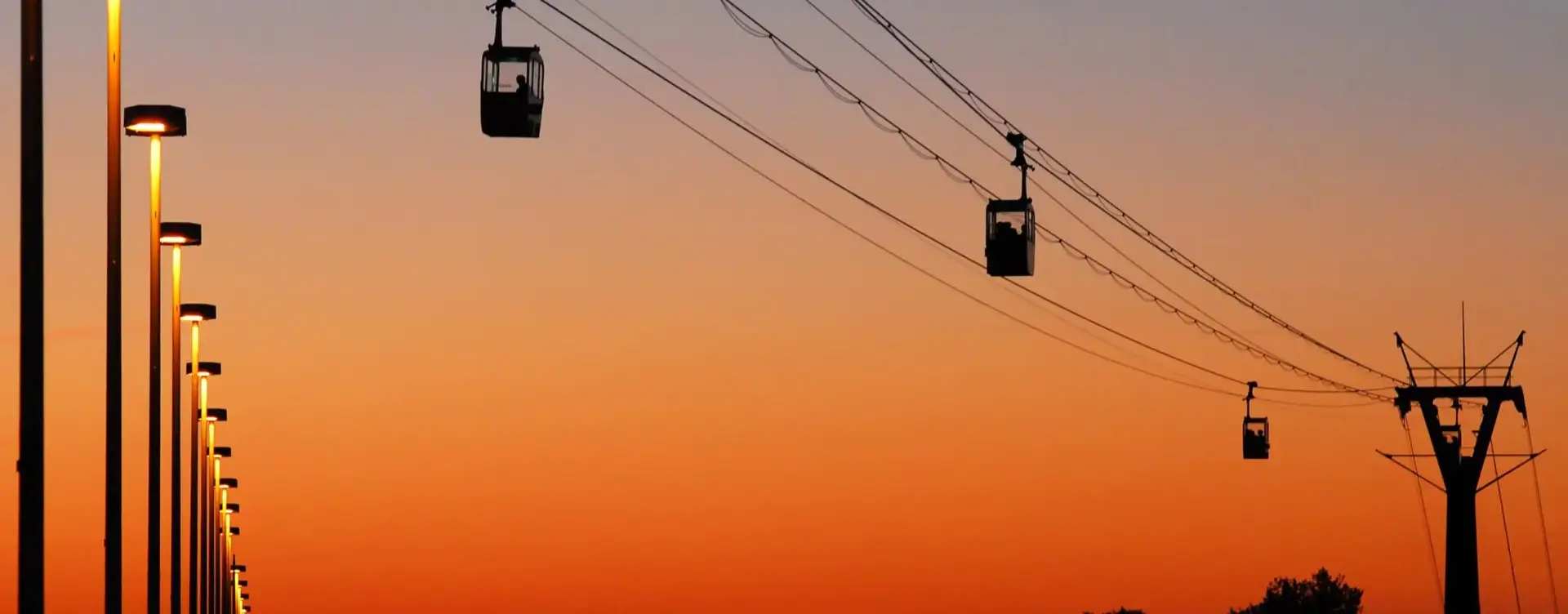 Seilbahnen auf Drähten heben sich vor einem orangefarbenen Abendhimmel ab, Straßenlaternen säumen die linke Seite.