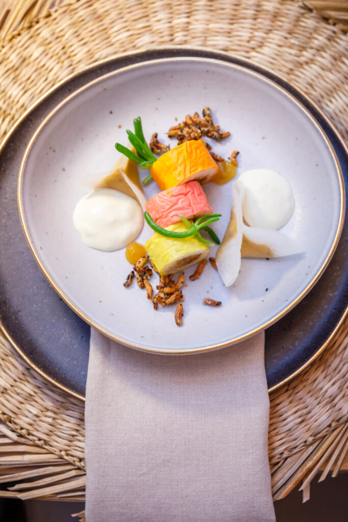 Elegantes Tellergericht mit bunten Würfeln, cremigen Klecksen, knuspriger Garnitur und Samen auf einem weißen Teller.