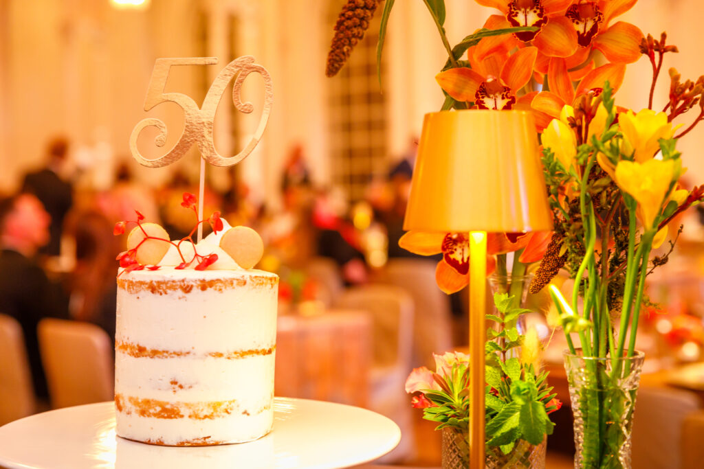 Zweistöckiger Zitronenkuchen mit goldener ‚50‘, umrahmt von Blumen in Orangetönen, 50. Jubiläumsfeier der GV-Praxis.