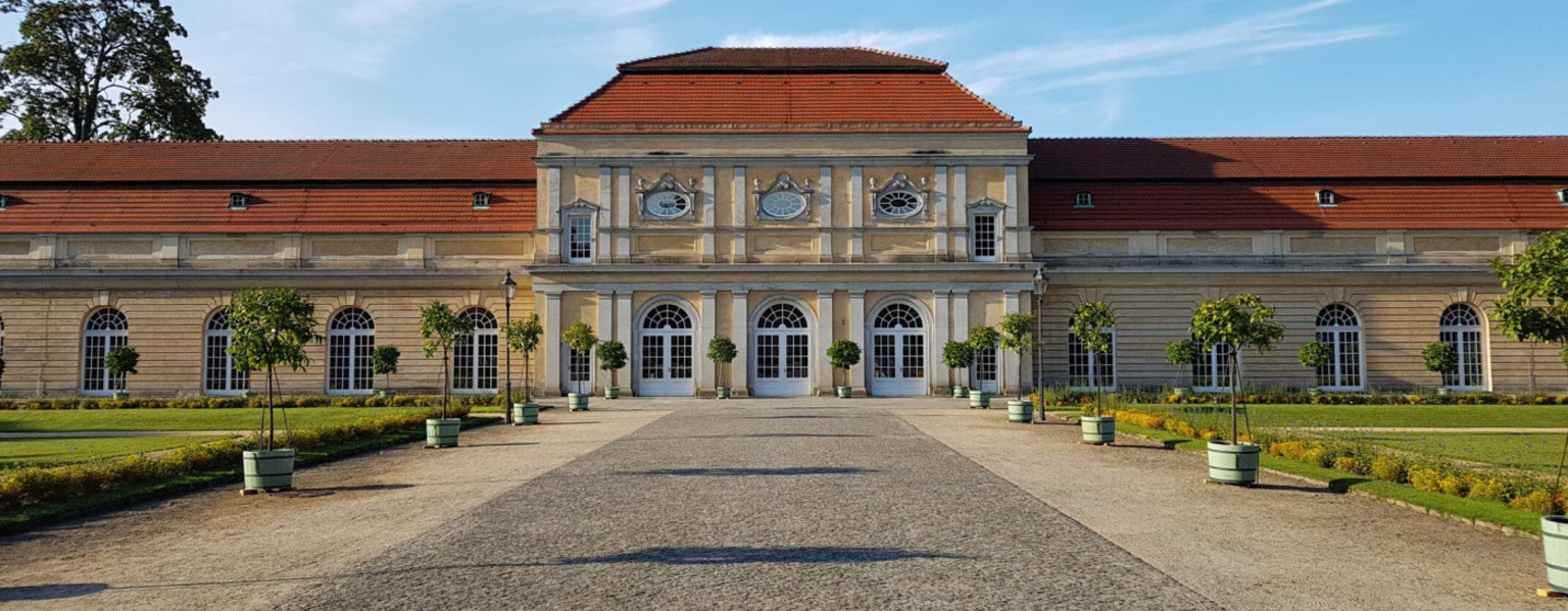 Ein prächtiges historisches Gebäude mit Bogenfenstern, einem roten Dach und einem von Bäumen gesäumten Weg, der zum Eingang führt – perfekt für Hochzeiten in der Orangerie Charlottenburg.