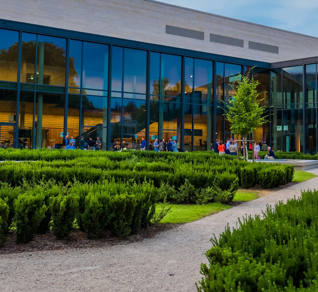 Ein moderner Glasbau mit Menschenansammlungen im Außenbereich und grünen Hecken im Vordergrund – ein idealer Ort, perfekt, wenn Sie Eventlocations in Köln mieten möchten.