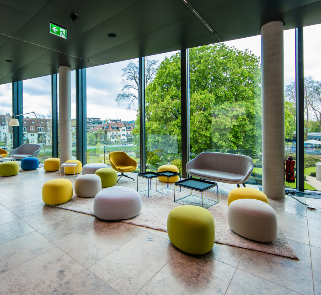 Moderne Lounge mit farbenfrohen Poufs, Sofas, großen Fenstern und Blick auf Bäume und Gebäude – perfekt für alle, die Eventlocations in Köln mieten möchten.