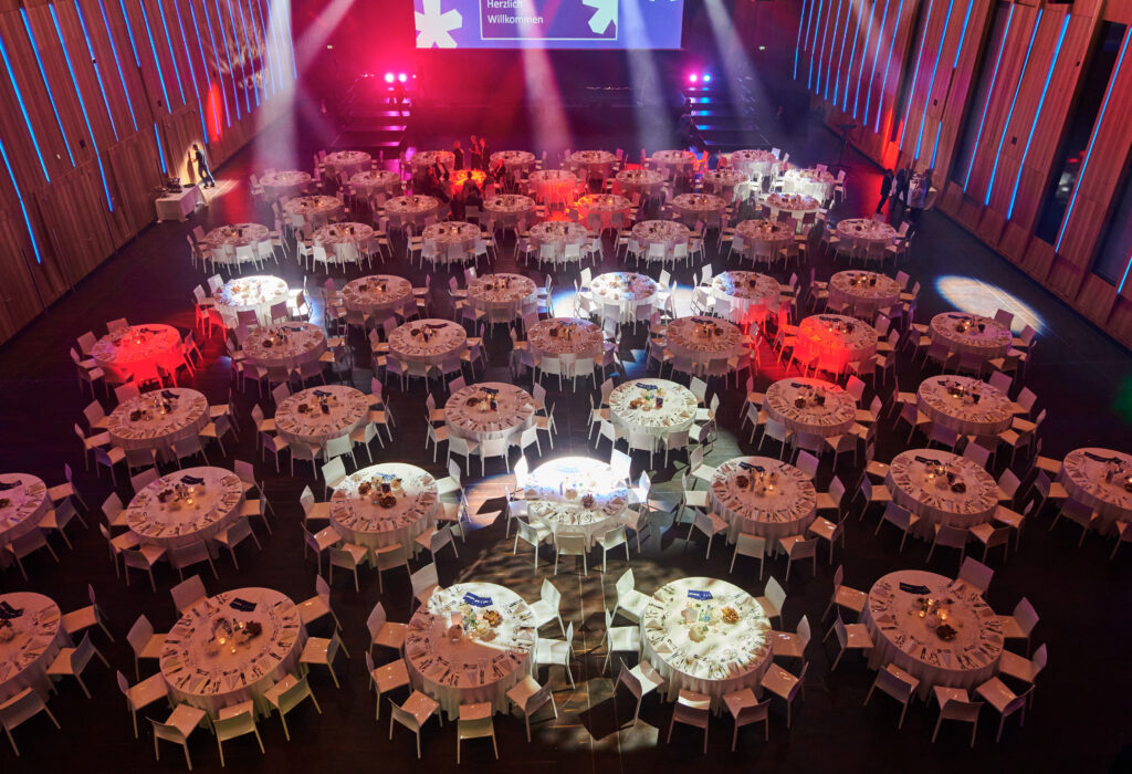 Ein großer Bankettsaal mit vielen runden Tischen, die für eine formelle Veranstaltung gedeckt und mit bunten Lichtern beleuchtet sind.