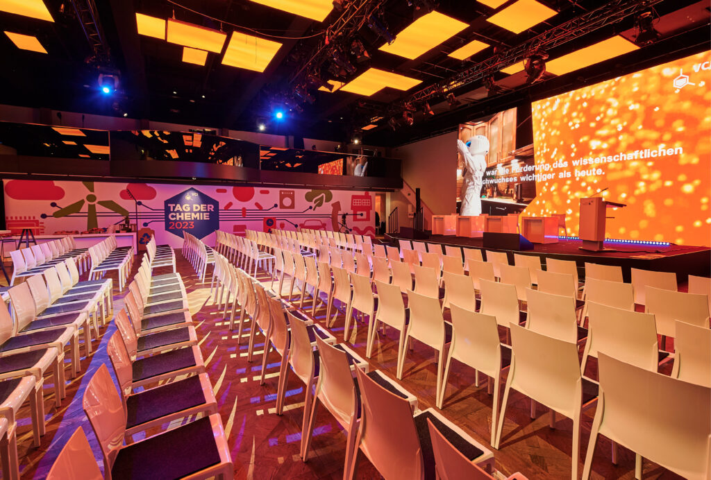 Palais Frankfurt - Reihen leerer Stühle stehen vor einer orange beleuchteten Bühne bei einer modernen Konferenzveranstaltung, dem Tag der Chemie 2023.