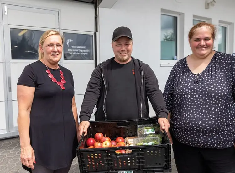 Drei Personen stehen lächelnd draußen, während eine eine Kiste mit Äpfeln und abgepackten Produkten hält.
