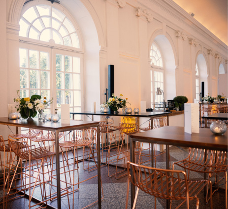 Eleganter Veranstaltungsraum mit hohen Tischen, Drahtstühlen, floralen Tischdekorationen und großen Bogenfenstern, die Licht hereinlassen – ideal für alle, die Eventlocations in Köln mieten möchten.