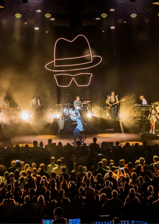 Eine Band tritt auf der Bühne auf, unter hellem Licht und mit einem neonfarbenen Hut und einer Brille darüber.