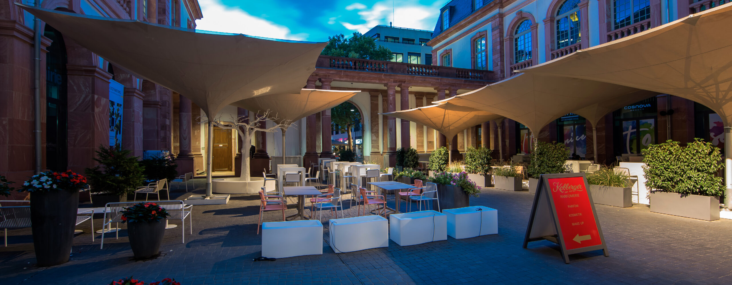 Palais Frankfurt - Café-Sitzbereich im Freien bei Einbruch der Dunkelheit mit modernen Zelten, Tischen, Stühlen, Pflanzen und einer roten Menü-Angabe.