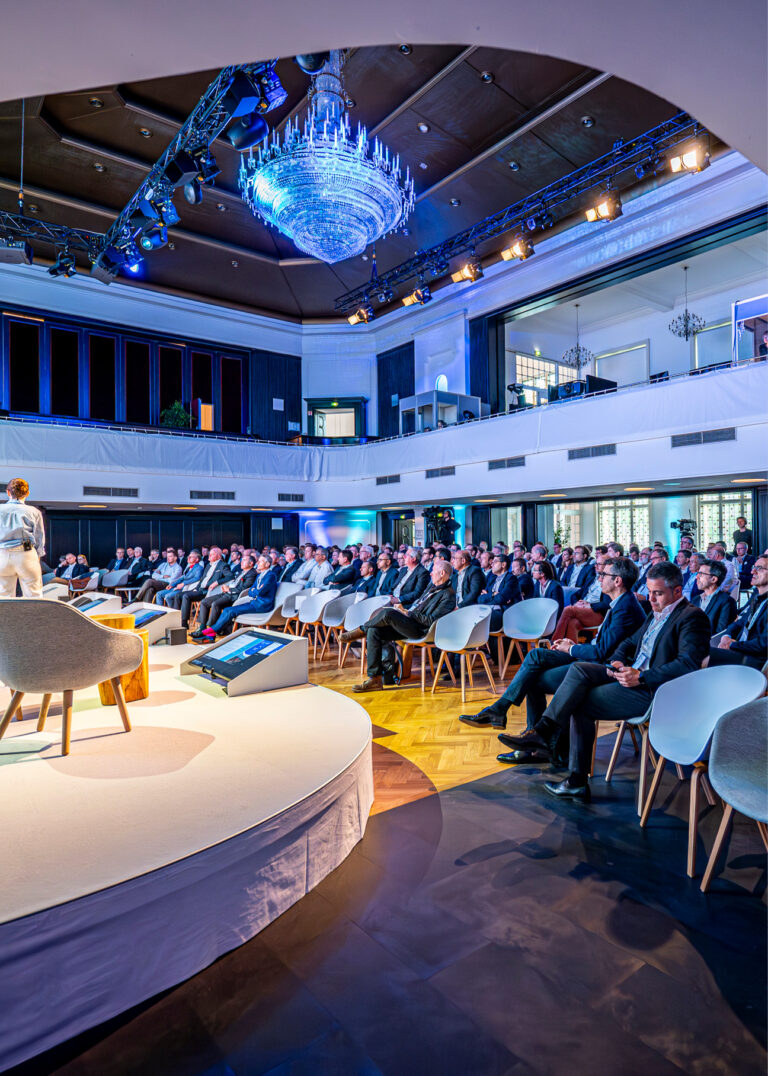 Das Publikum sitzt in einem modernen Auditorium und beobachtet einen Redner auf einer hell erleuchteten Bühne mit einem großen Kronleuchter darüber.
