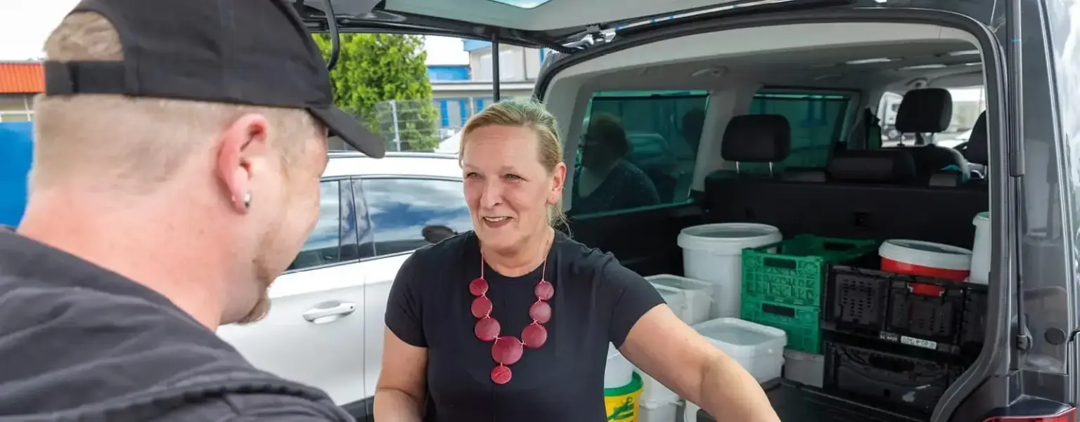 Eine Frau steht neben einem offenen Kofferraum und unterhält sich mit einem Mann; im Auto befinden sich Eimer und Kisten.