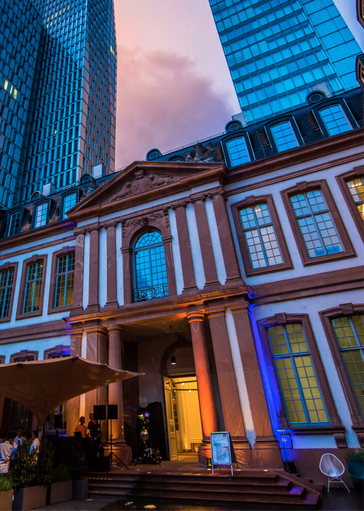 Palais Frankfurt - Historisches Gebäude mit Säulen und großen Fenstern, das in der Dämmerung beleuchtet wird und von modernen Glaswolkenkratzern flankiert wird.