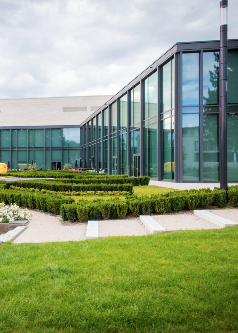 Modernes Glasgebäude mit gepflegten Hecken, grünem Rasen und bewölktem Himmel.