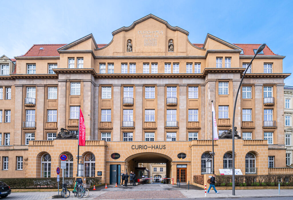 Das Curio-Haus, ein historisches beiges Gebäude in Hamburg, besticht durch Säulen, Statuen und rote Dachziegel – ein zeitloser Charme, der es mit den Top-Eventlocations in Köln aufnehmen kann, die für besondere Anlässe gemietet werden.