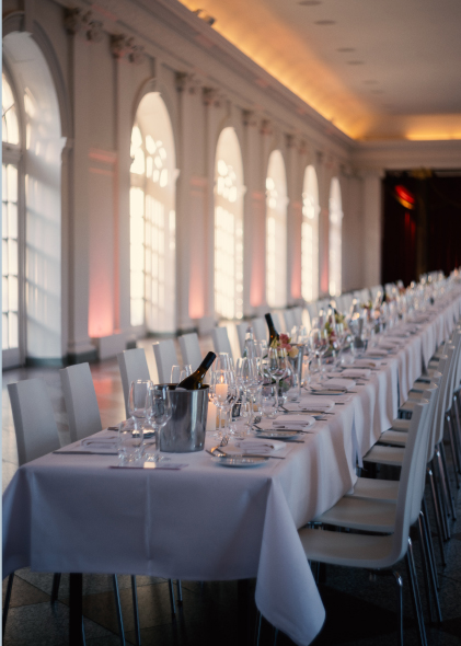 Ein langer, elegant gedeckter Esstisch für eine Veranstaltung in einem hellen Saal mit großen Bogenfenstern in der Orangerie Charlottenburg für Hochzeiten.