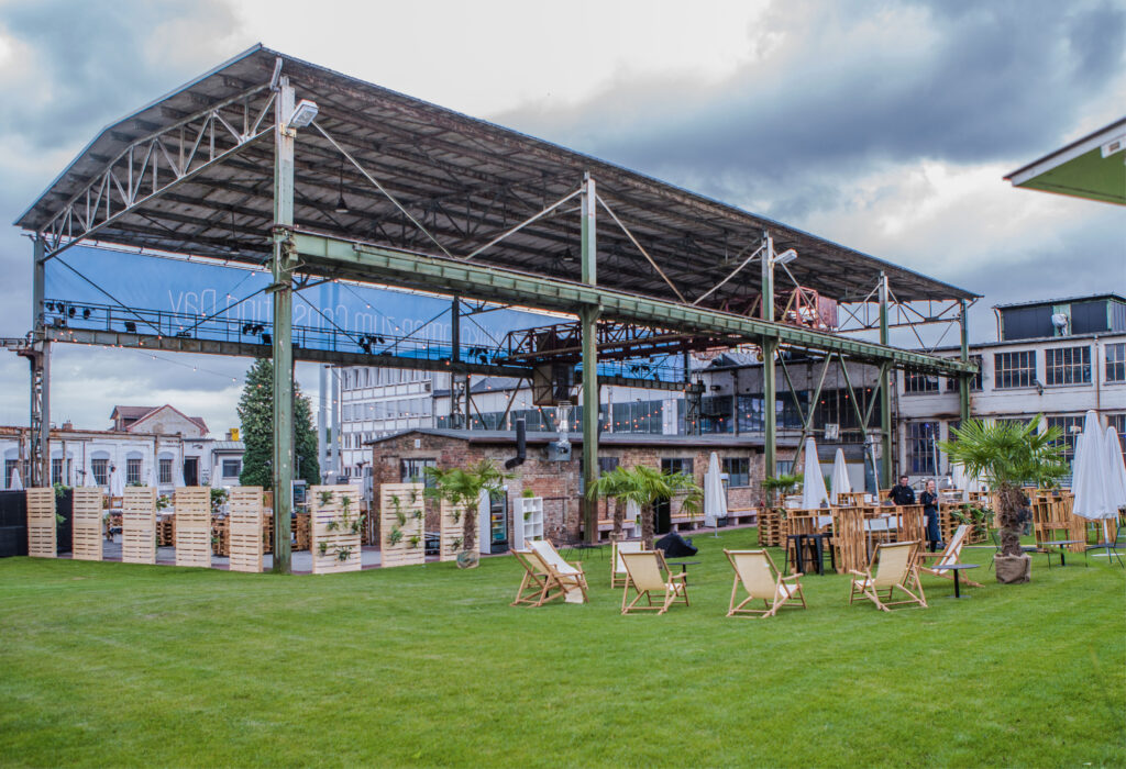 Veranstaltungsfläche im Freien in Fredenhagen als Veranstaltungsort mit Gartenstühlen, Palmen und einer großen offenen Metallkonstruktion unter einem bewölkten Himmel.