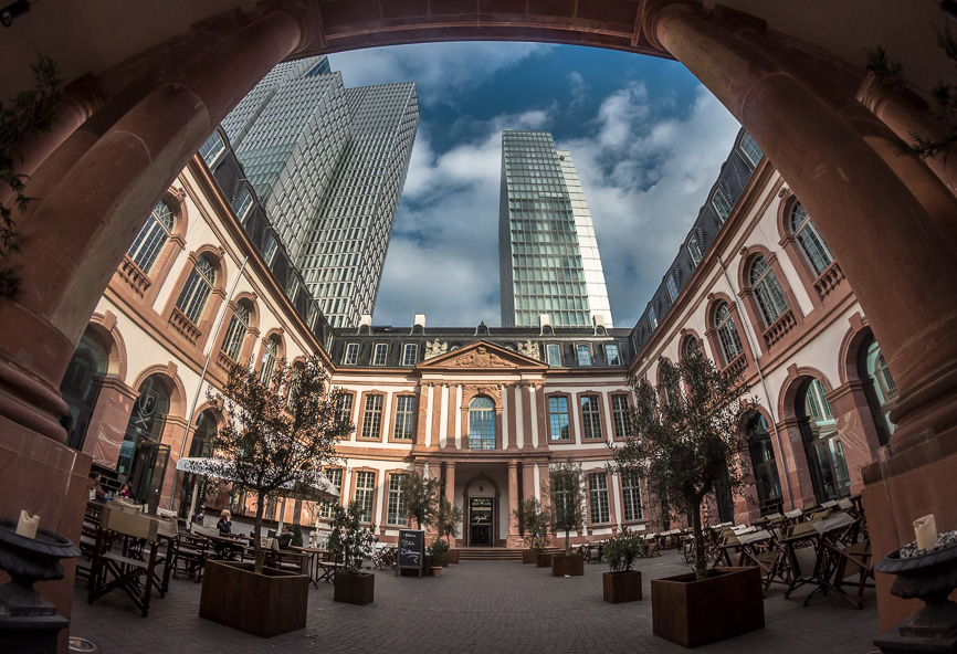 Ein historischer Innenhof mit Sitzgelegenheiten im Freien, eingerahmt von Torbögen und modernen Wolkenkratzern – ein einzigartiger Ort für alle.