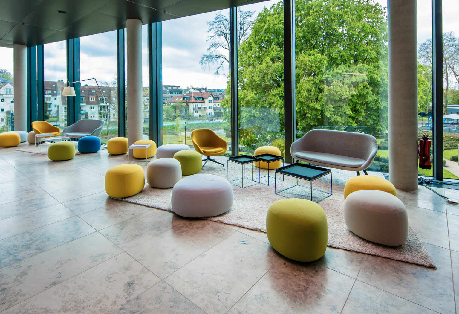 Moderne Lounge mit farbenfrohen Poufs, Stühlen und großen Fenstern mit Blick auf Bäume und die Stadtlandschaft.