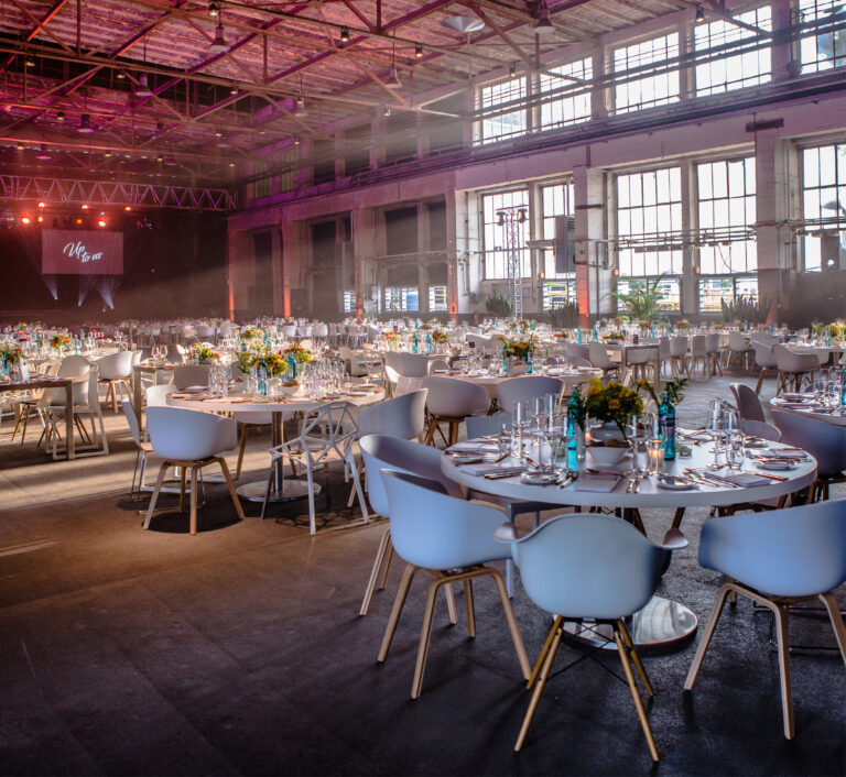 Moderner Bankettsaal mit runden Tischen, weißen Stühlen, floralen Tischdekorationen und großen Fenstern mit natürlichem Licht – ideal für alle, die Eventlocations in Köln für besondere Anlässe mieten möchten.