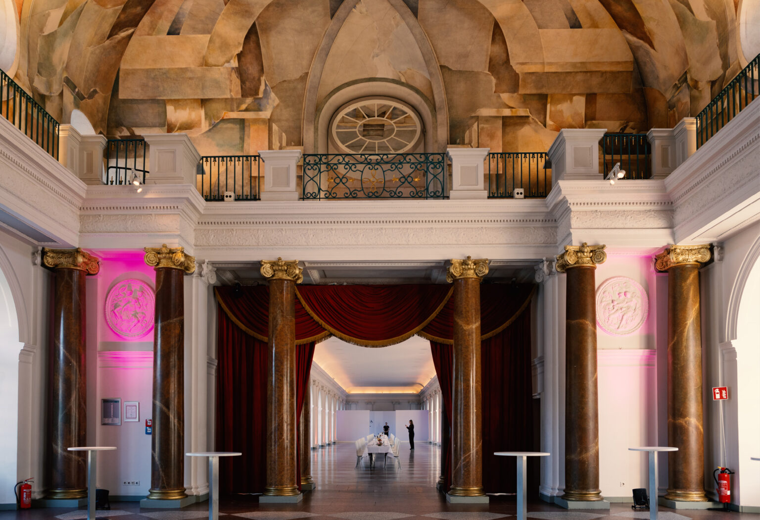 Großer Saal mit Marmorsäulen, verzierter Decke, roten Vorhängen und einem langen Korridor mit Tischen und rosafarbener Beleuchtung – perfekt geeignet als Orangerie Charlottenburg für Hochzeiten.