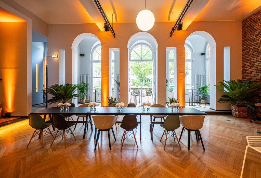 Curio-Haus - Moderner Tagungsraum mit langem Tisch, Stühlen, Pflanzen, großen Fenstern und warmer Beleuchtung.