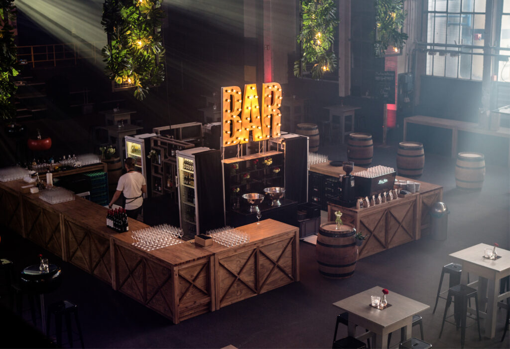 Eine moderne Bar mit Holztheken, Fässern und einem großen beleuchteten BAR-Schild in einem Raum im Industriestil – perfekt für alle, die Eventlocations in Köln mieten möchten.