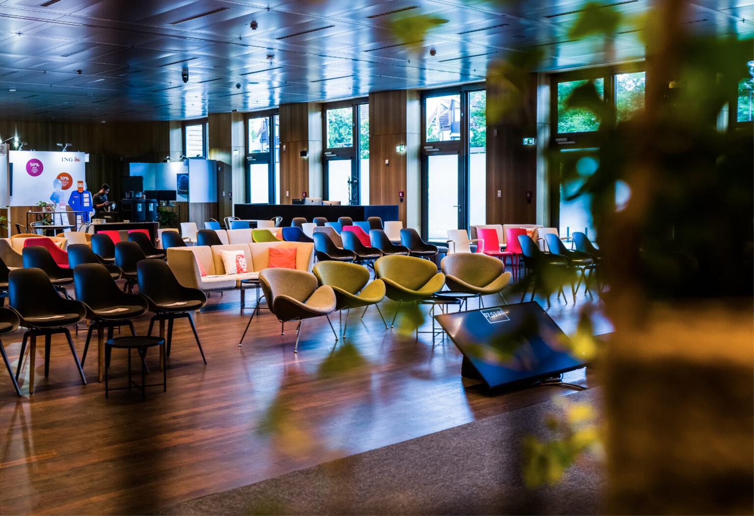 Moderner Veranstaltungsraum mit bunten, in Reihen angeordneten Stühlen, großen Fenstern und heller Beleuchtung.