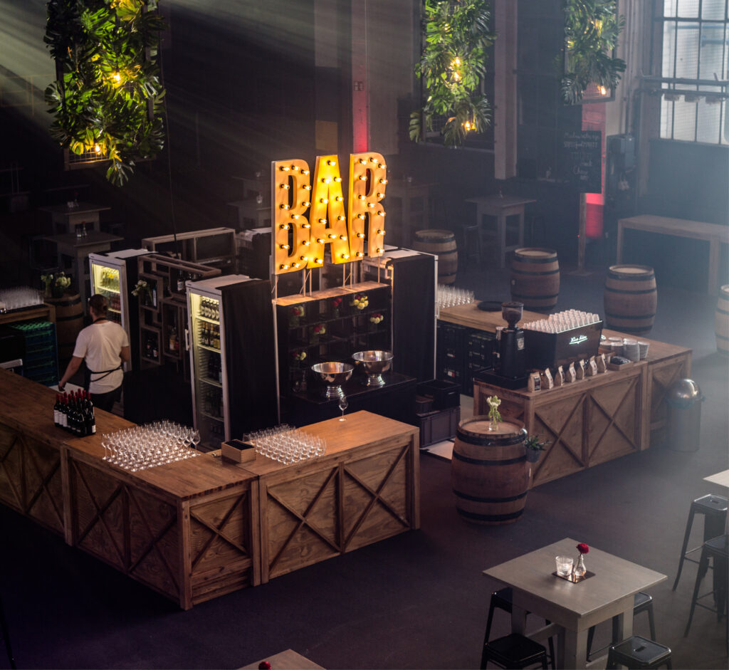 Großzügige Indoor-Bar mit beleuchtetem BAR-Schild, Holztheken, Weingläsern und üppigem Grün.