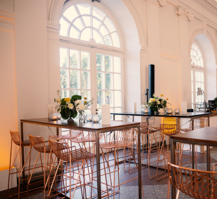 Modernes Café mit hohen Tischen, Kupferstühlen und großen Bogenfenstern, die natürliches Licht hereinlassen – perfekt für alle, die die elegante Atmosphäre der Orangerie Charlottenburg für Hochzeiten lieben.