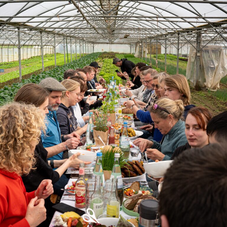 Eine große Gruppe von Menschen isst gemeinsam an einem langen Tisch in einem von Pflanzen umgebenen Gewächshaus und unterstreicht so die Nachhaltigkeit im Catering in einem üppigen, umweltfreundlichen Ambiente.