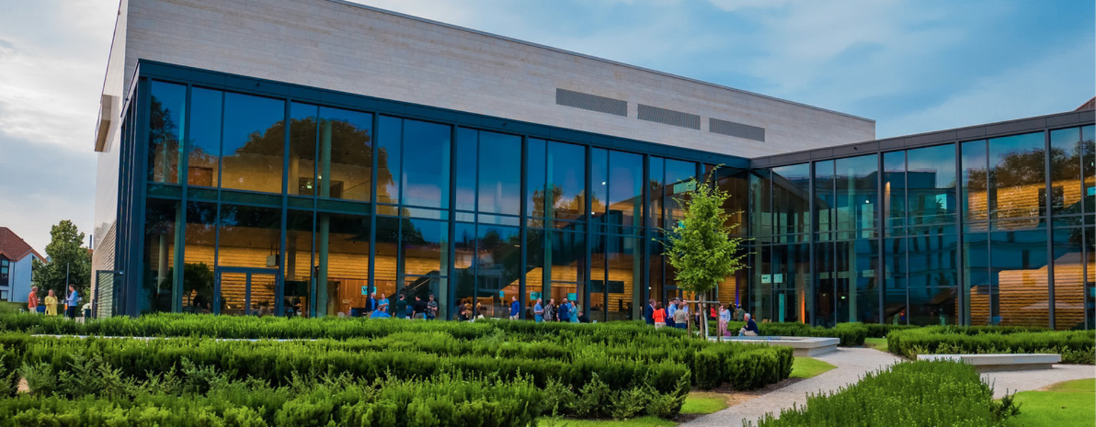 Modernes Glasgebäude mit Menschen darin, umgeben von grünen Büschen und Wegen an einem bewölkten Tag – perfekt für Versammlungen, die von Vilco Catering Services unterstützt werden.