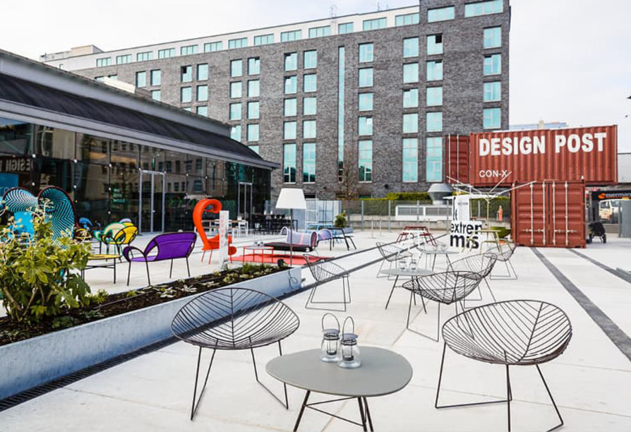 Moderne Außenterrasse mit bunten Stühlen, Tischen und einem roten DESIGN POST-Container im Hintergrund.