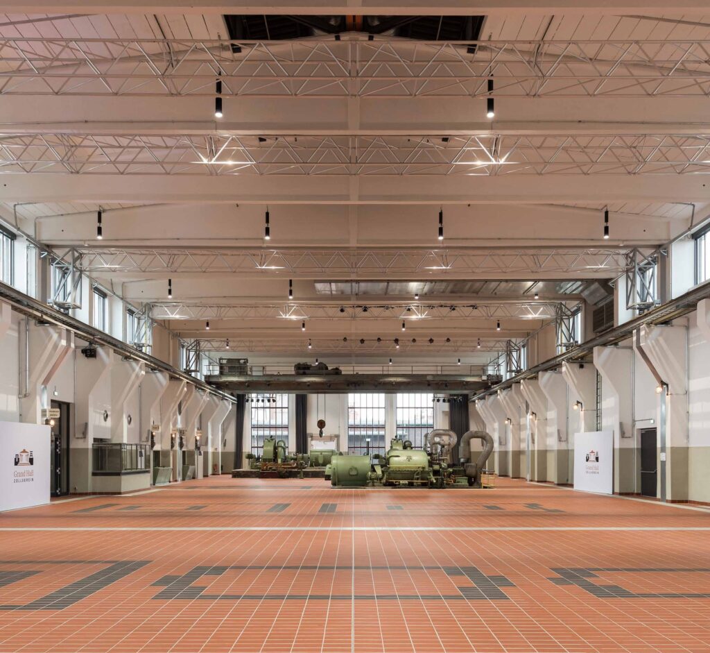 Geräumige Industriehalle mit rotem Fliesenboden und großen grünen Maschinen am anderen Ende – ideal für alle, die Eventlocations in Köln mieten möchten.