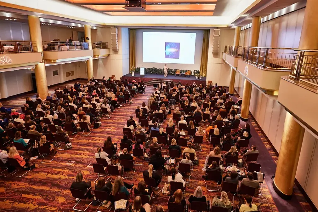 Ein großes Publikum sitzt in einem Konferenzsaal und verfolgt, wie eine Rednerin auf einer Bühne mit einem Bildschirm die aktuellsten Neuigkeiten des Frauenforum Foodservice präsentiert.